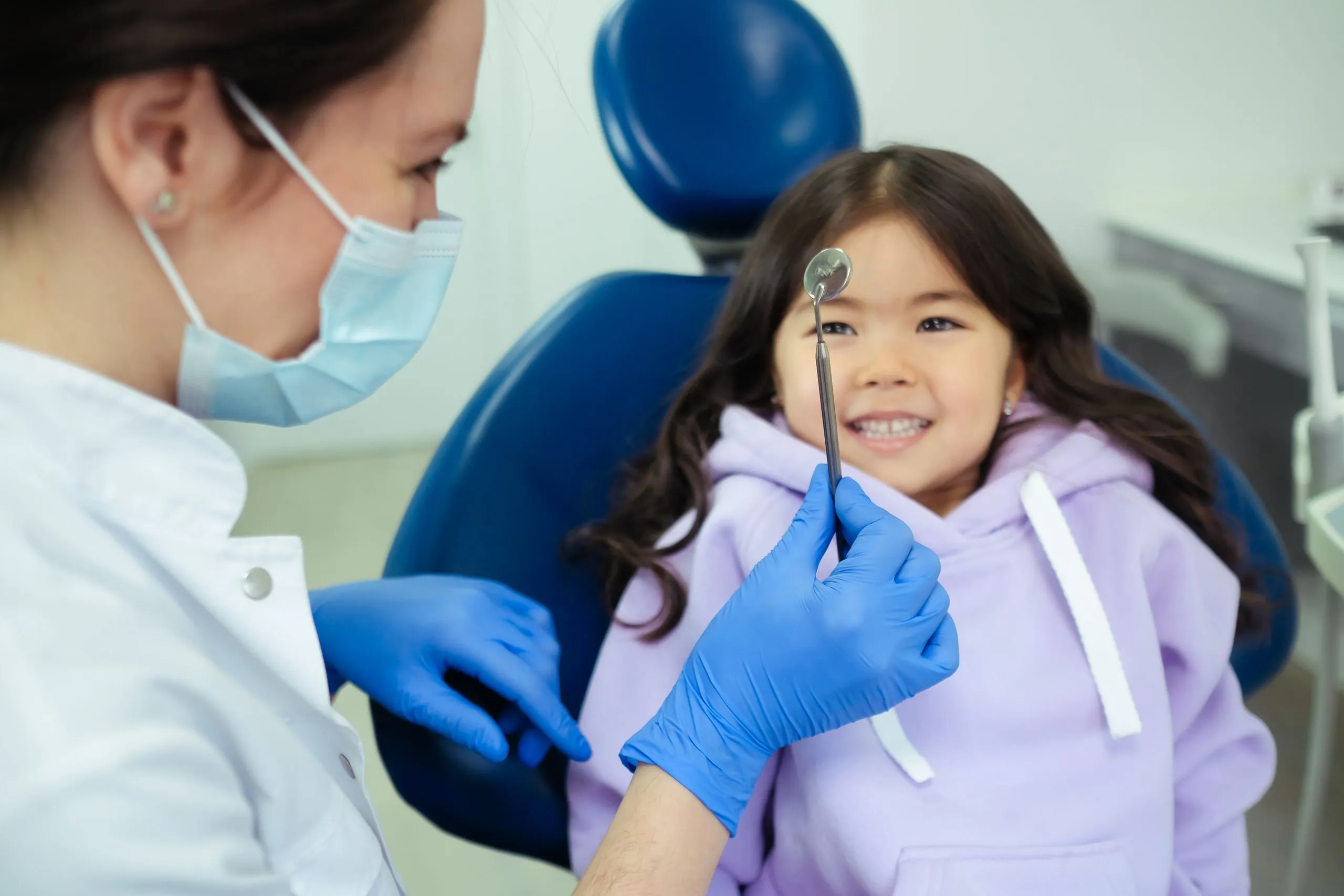 Une dentiste présente un miroir d’examen à une enfant souriante installée au fauteuil.