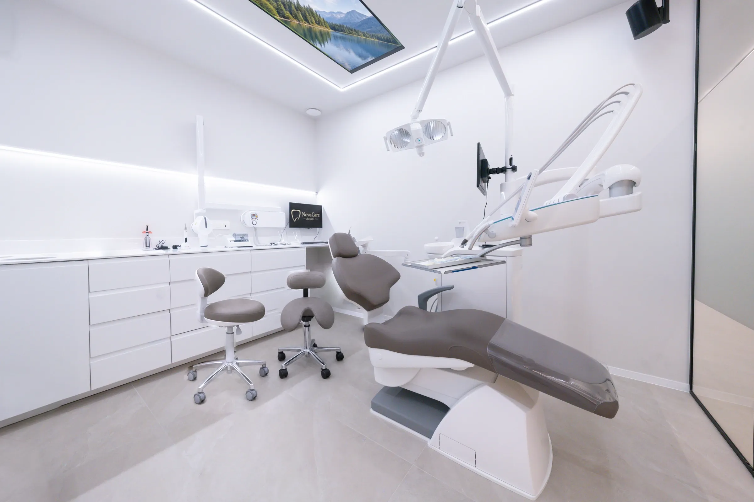 La salle de soins de NovaCare Dental à Jette, avec son fauteuil gris, sa double lampe et son écran au plafond.