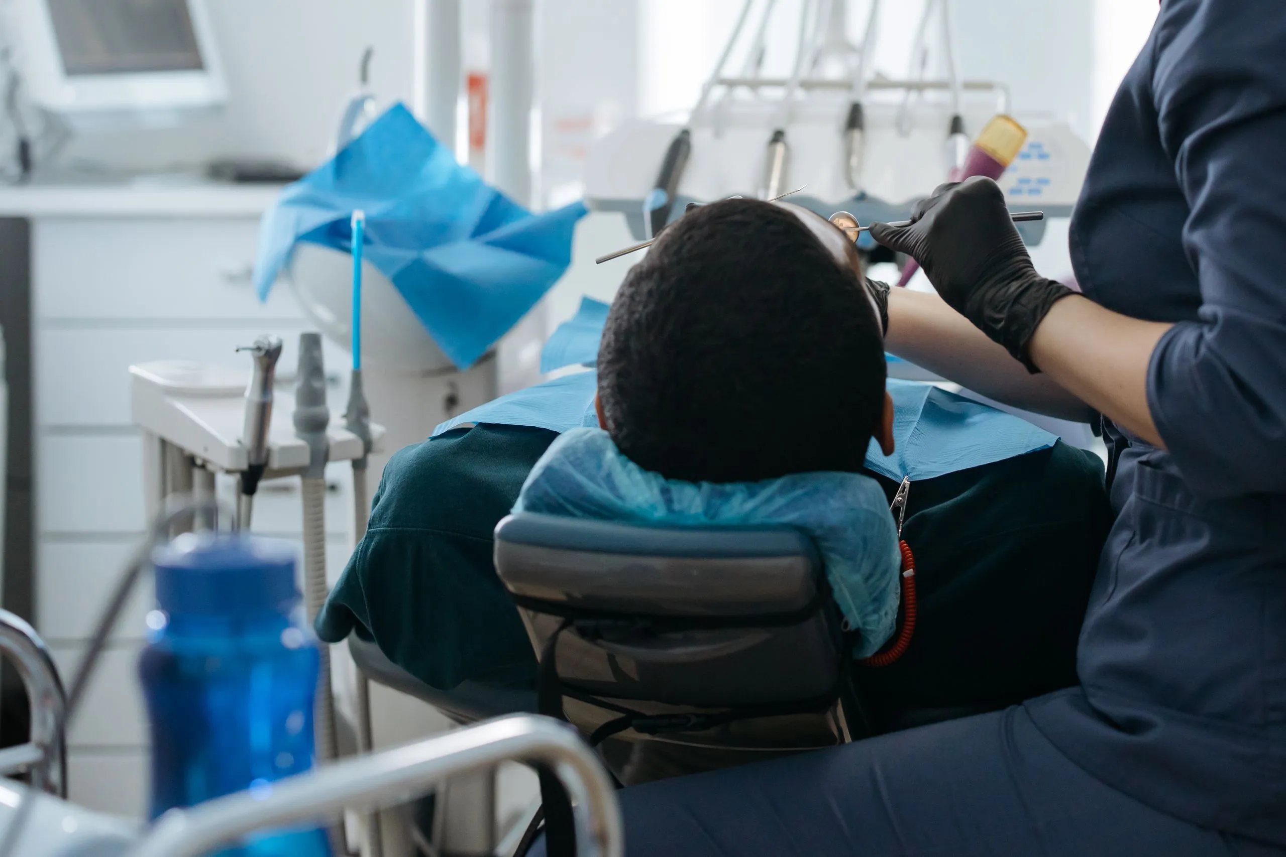 Vue de dos d’une personne au fauteuil pendant un soin dentaire, avec les mains gantées du praticien à ses côtés.